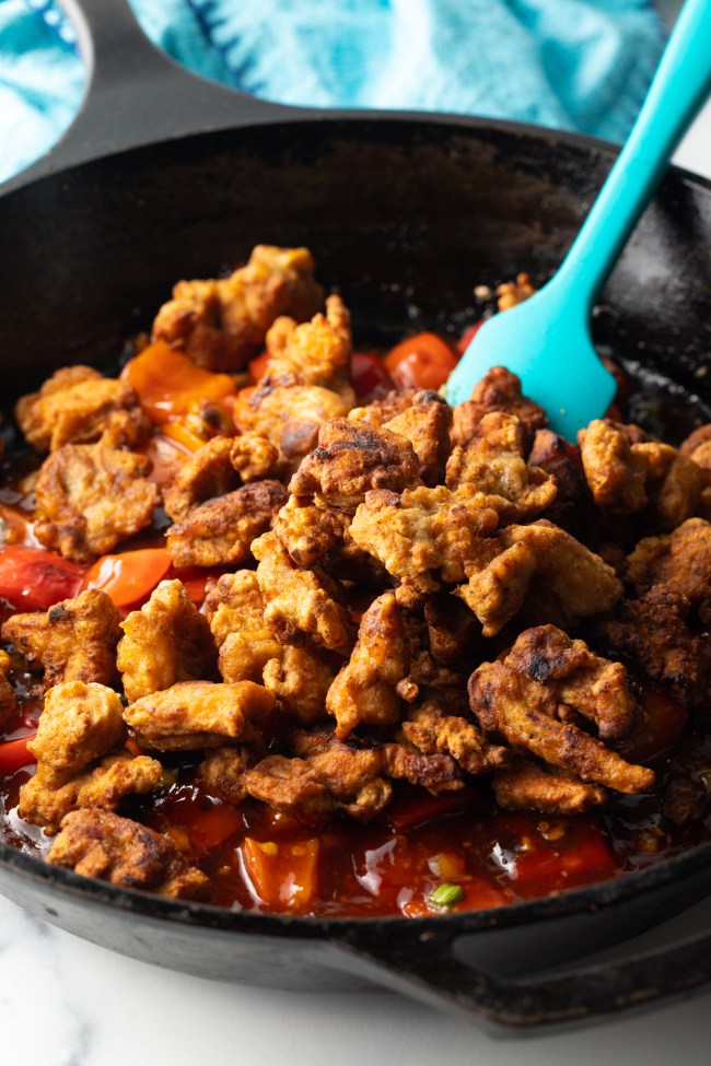 Blue spatula stirring chicken pieces and red peppers in a skillet.
