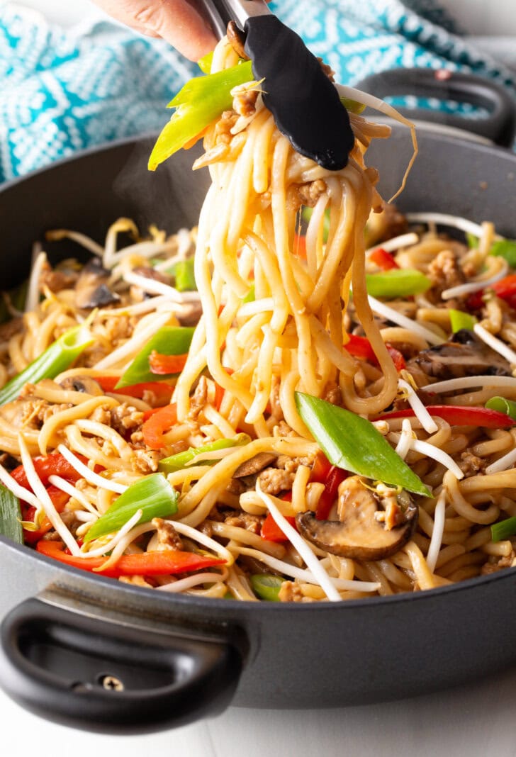 Tongs grabbing a serving of udon noodles with veggies and chicken.