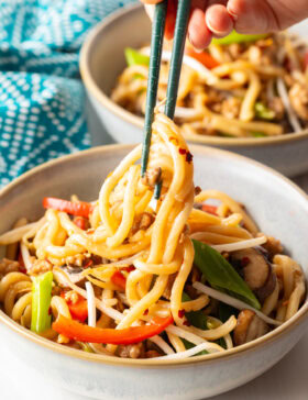 Hand with chopsticks grabbing some noodles from the bowl of stir fried vegetables and chicken.