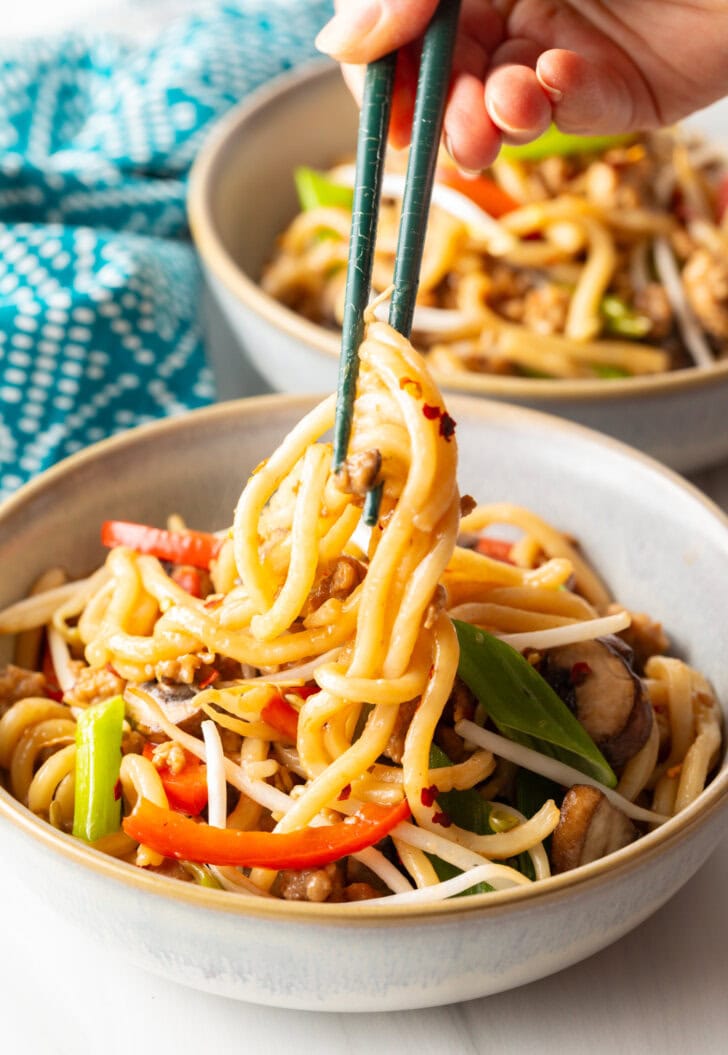 Hand with chopsticks grabbing some noodles from the bowl of stir fried vegetables and chicken.