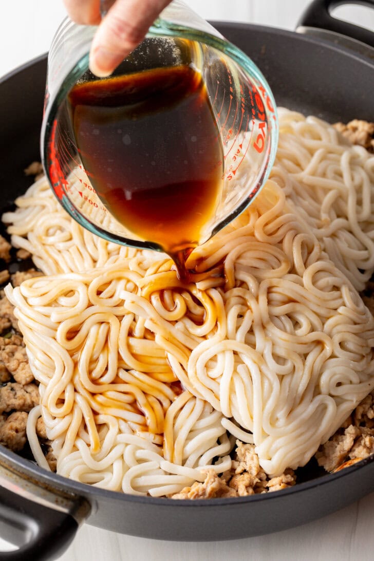 Pouring stir fry sauce over udon noodles in a large skillet.