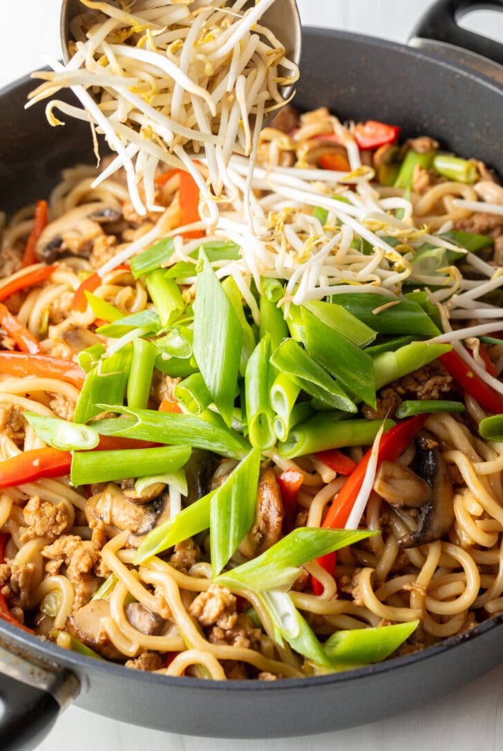 Close view skillet with yaki udon noodles, adding bean sprouts and the dish is topped with sliced green scallions