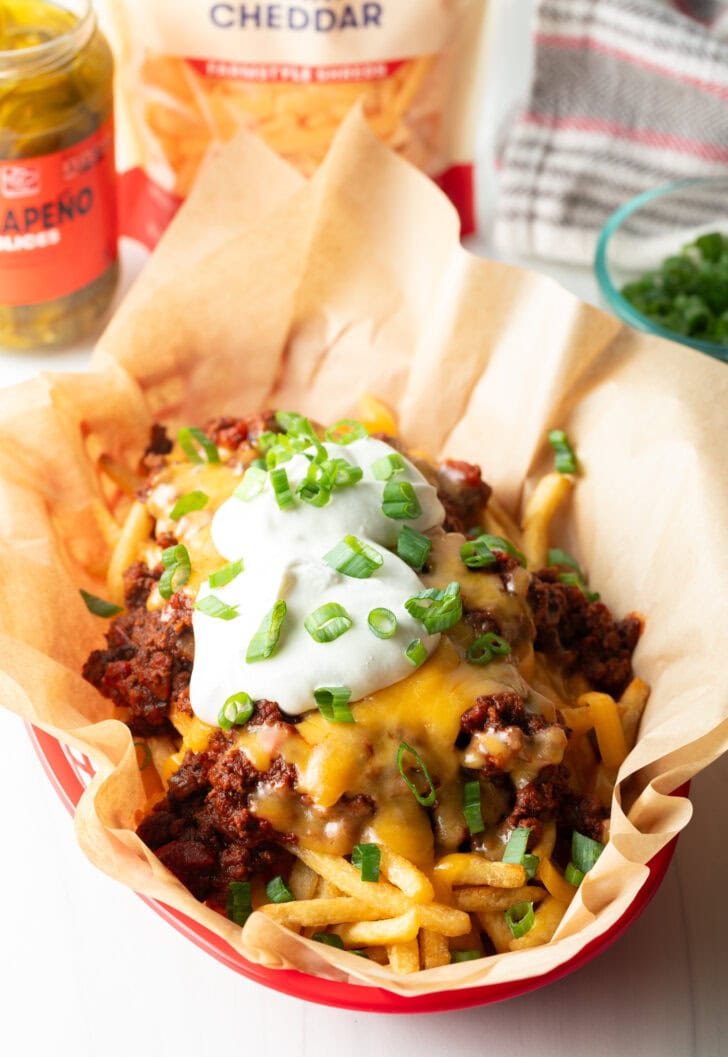 Basket with parchment paper, filled with crispy fries topped with chili and melted cheddar cheese, sour cream, and chopped green onions.