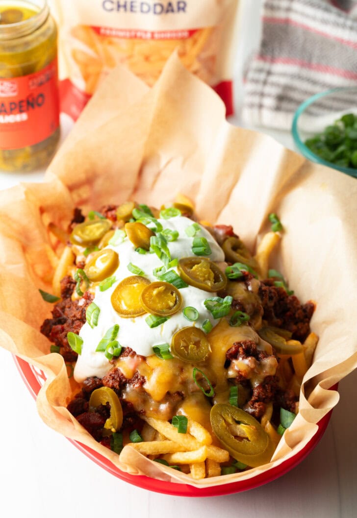 Basket with parchment paper, filled with crispy fries topped with chili and melted cheddar cheese, sour cream, chopped green onions, and jalapeno slices.