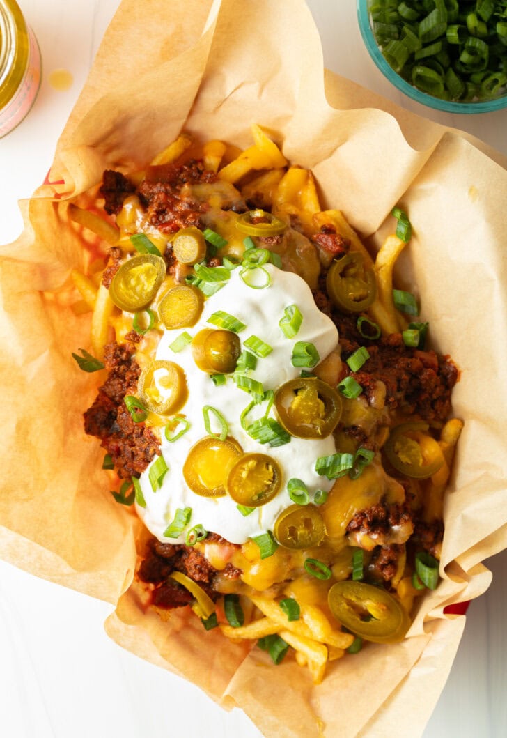 Basket with parchment paper, filled with crispy fries topped with chili and melted cheddar cheese, sour cream, chopped green onions, and jalapeno slices.