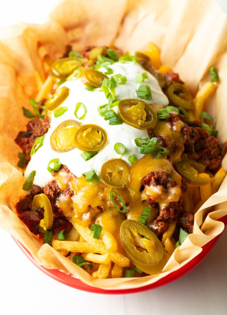 Basket with parchment paper, filled with crispy fries topped with chili and melted cheddar cheese, sour cream, chopped green onions, and jalapeno slices.