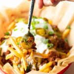 Basket with parchment paper, filled with crispy fries topped with chili and melted cheddar cheese, sour cream, chopped green onions, and jalapeno slices. A hand with forkful of fries is showing to camera.