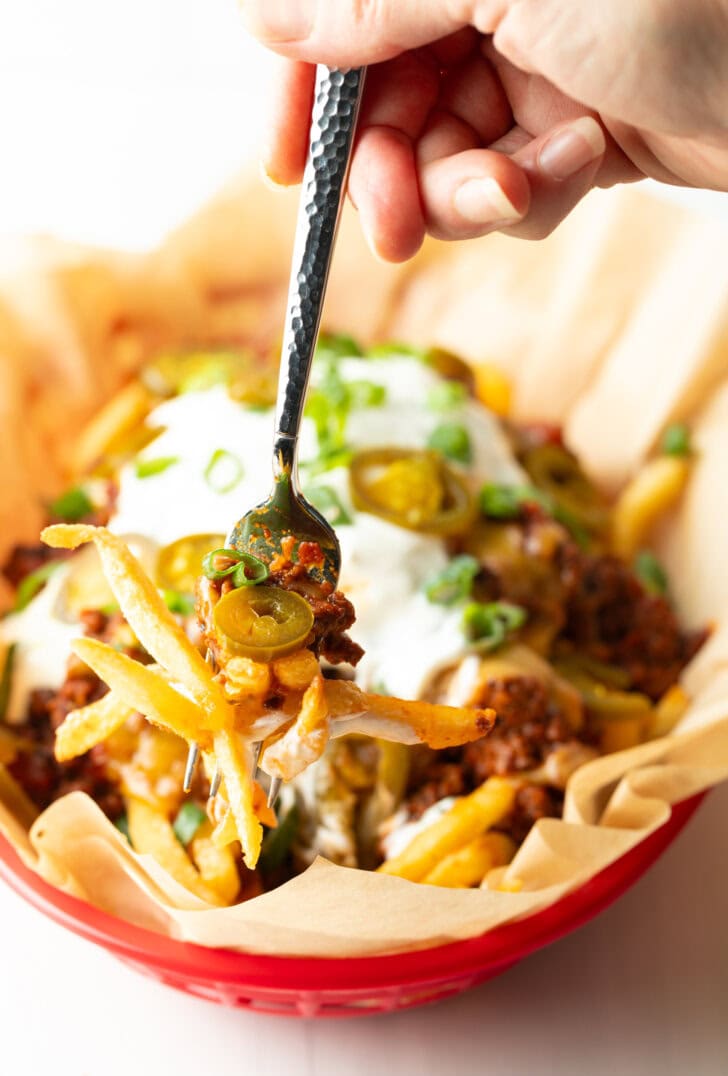 Basket with parchment paper, filled with crispy fries topped with chili and melted cheddar cheese, sour cream, chopped green onions, and jalapeno slices. A hand with forkful of fries is showing to camera.