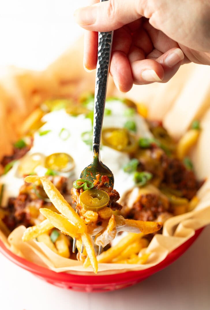 Basket with parchment paper, filled with crispy fries topped with chili and melted cheddar cheese, sour cream, chopped green onions, and jalapeno slices. A hand with forkful of fries is showing to camera.