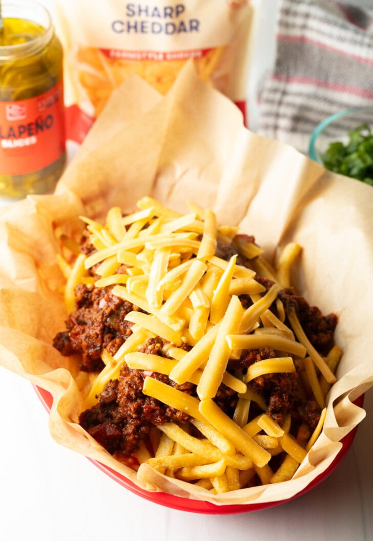 Basket with parchment paper, filled with crispy fries topped with chili and shredded cheddar cheese.