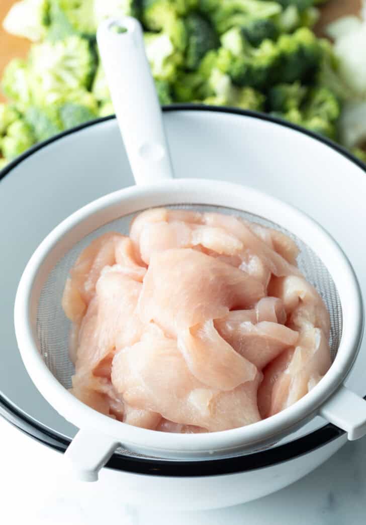 Draining pieces of raw chicken in a colander over a white bowl.