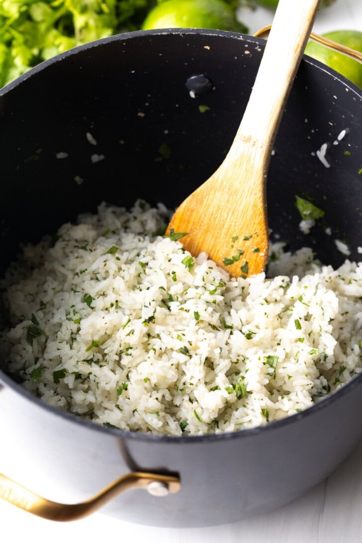 Wooden spatula stirring fluffy white rice with chopped cilantro in a large cooking pot.