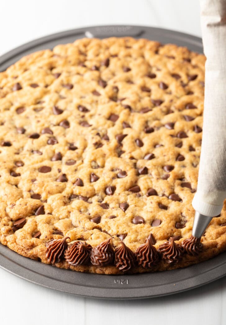 Piping chocolate frosting along the edge of the cookie cake.