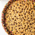 Top down view large chocolate chip cookie cake. The outside edge is piped with chocolate frosting and covered with rainbow sprinkles.