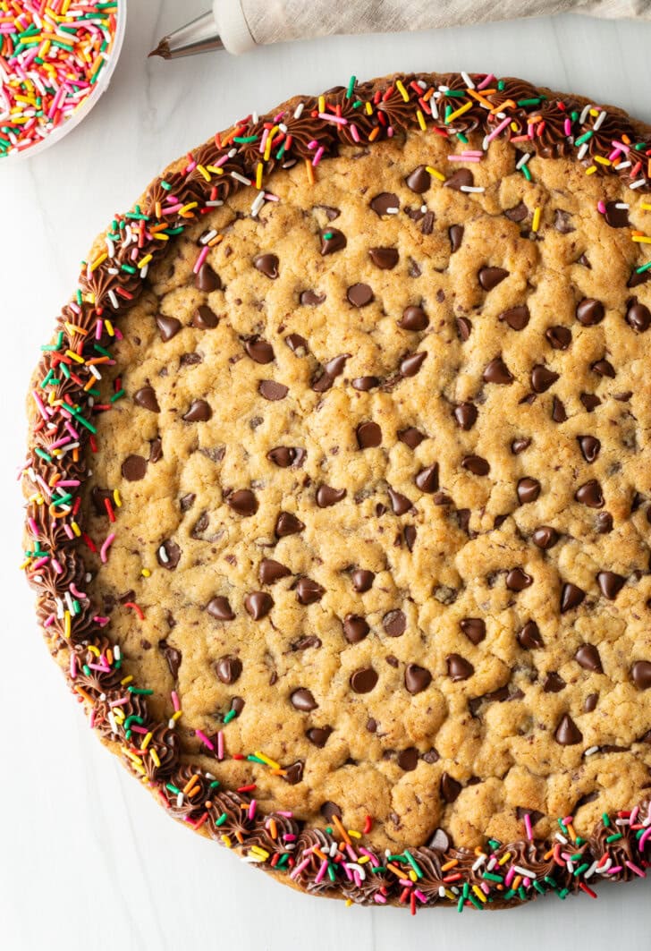 Top down view large chocolate chip cookie cake. The outside edge is piped with chocolate frosting and covered with rainbow sprinkles.