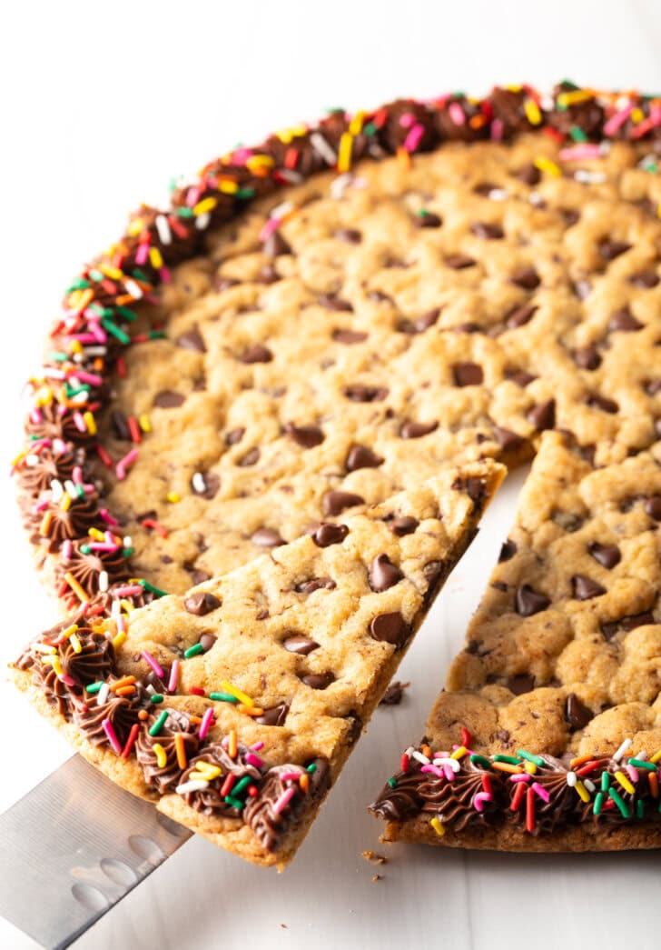 Large chocolate chip cookie cake. The outside edge is piped with chocolate frosting and covered with rainbow sprinkles. A cake cutter is taking a wedge slice from the cake.