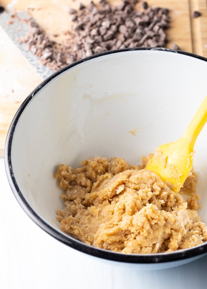 White mixing bowl with cookie dough ingredients, stirred by a yellow spatula.