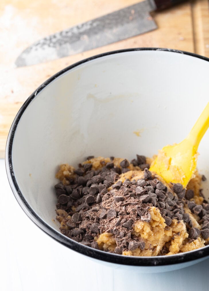 White mixing bowl with cookie dough ingredients, stirred by a yellow spatula.