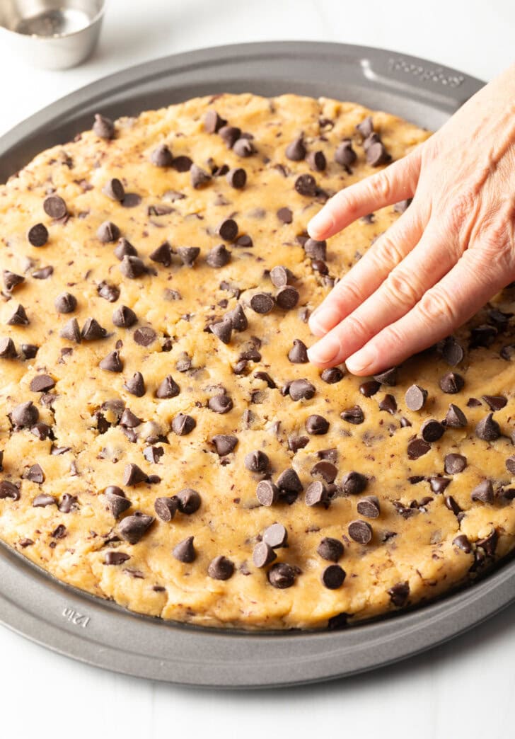 Large chocolate chip cookie cake, hand spreading dough out in a pizza pan.