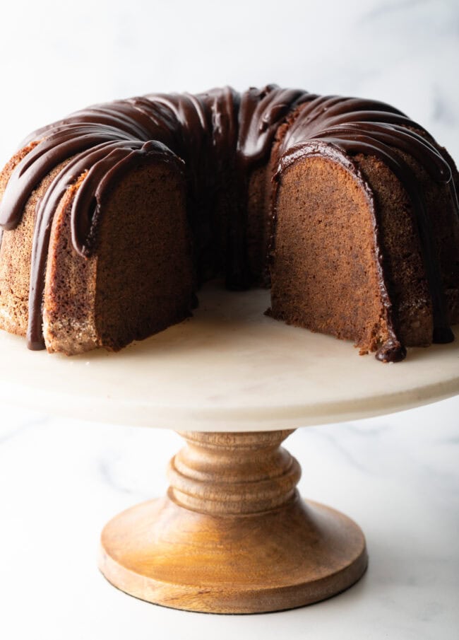 Chocolate sour cream bundt cake with a thick chocolate glaze drizzled on top. The cake is on a white stand and has a portion removed to show the tender inside crumb.