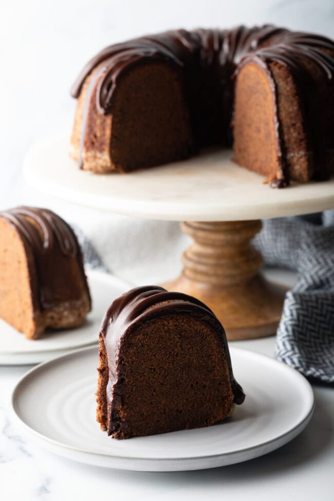 Chocolate Glaze for Pound Cake - Thick slice of chocolate sour cream pound cake with drizzle of glaze on a white plate. The full cake is on a white cake stand in the background.