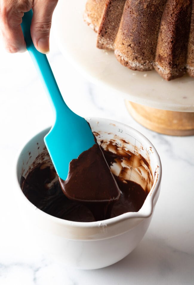 White measuring pitcher, hand with blue spatula is stirring a rich chocolate glaze.