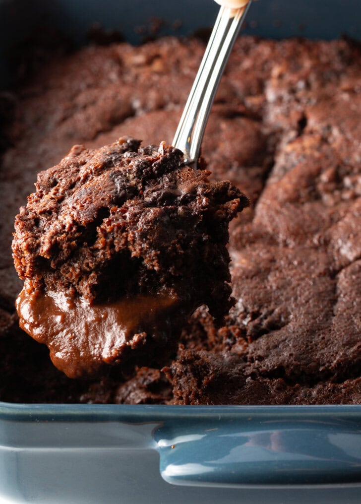 Hand using a metal spoon to scoop a serving of pudding cake from the baking pan.