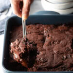 Hand using a metal spoon to scoop a serving of pudding cake from the baking pan.