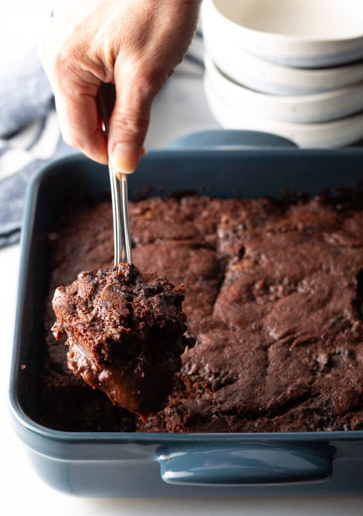 Hand using a metal spoon to scoop a serving of pudding cake from the baking pan.