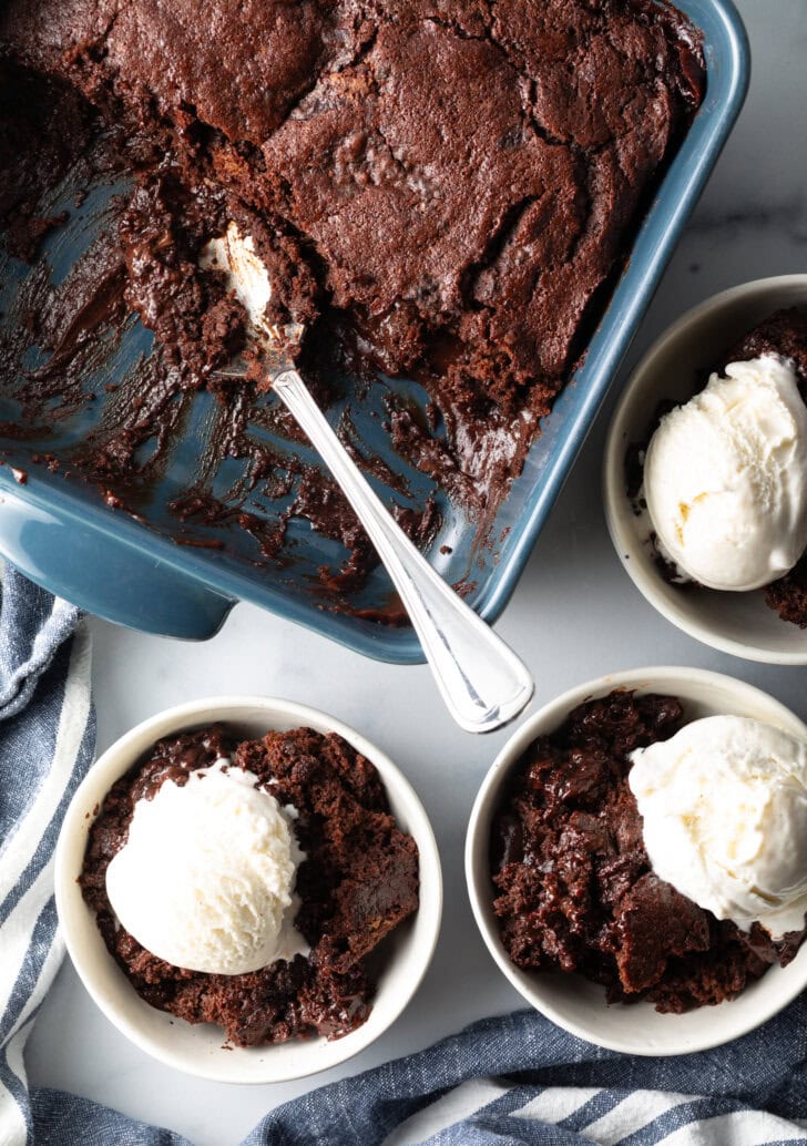 3 white bowls with servings of chocolate lava cake and a scoop of vanilla ice cream. Bowls are next to a baking pan with pudding cake and a metal spoon.