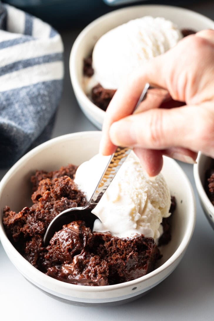 White bowl with serving of chocolate lava cake and a scoop of vanilla ice cream. A hand with a metal spoon is taking a scoop from the bowl.