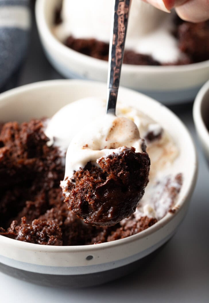 White bowl with serving of chocolate lava cake and a scoop of vanilla ice cream. A hand with a metal spoon is taking a scoop from the bowl.