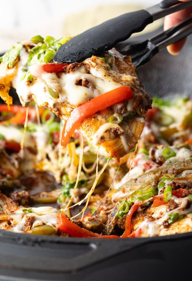 Choripollo in a black skillet, topped with cheese and green onions. A hand with tongs is taking a bit of the peppers and chicken.