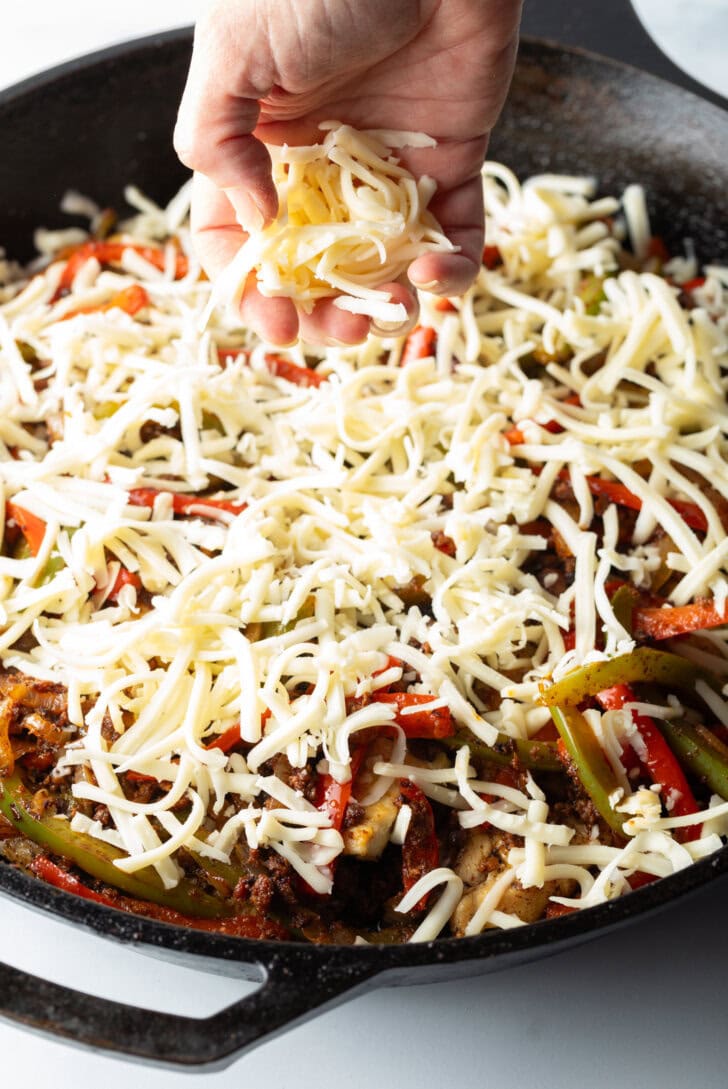 Hand adding shredded cheese on top of the chicken and pepper in the black skillet.