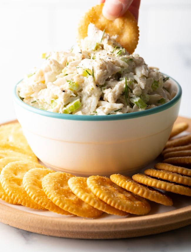 Hand holding a cracker that is scooping crab salad from a bowl. 