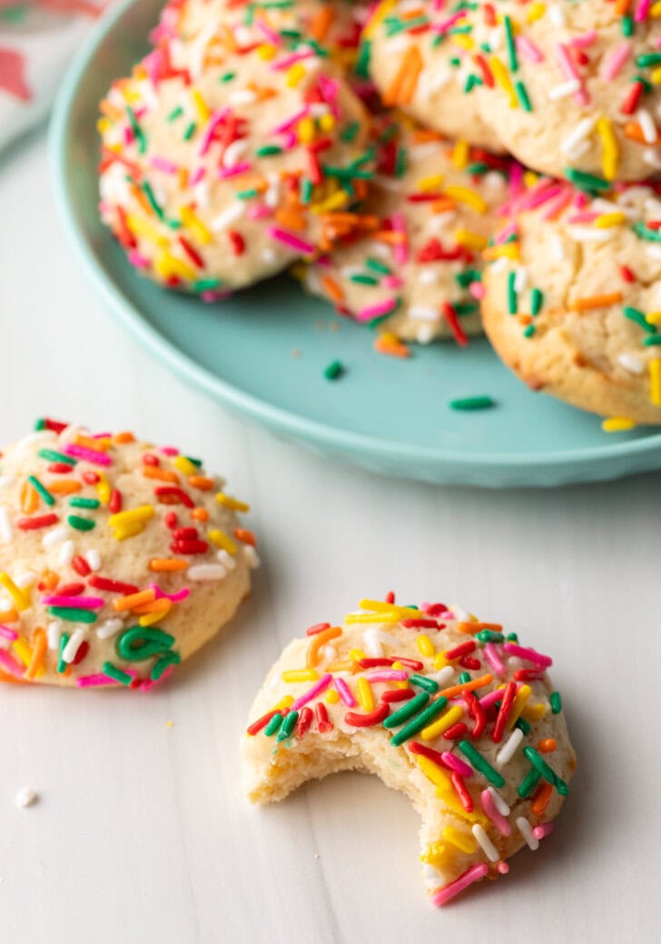 Cookie with sprinkles and a bit taken out is close to camera. Another cookie with no bite is off to the side. In the background is a plate piled up with cream cheese cookies.