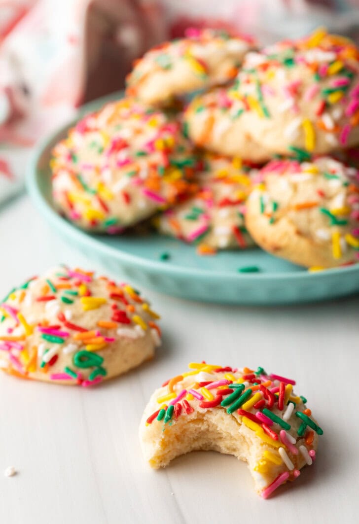 Cookie with sprinkles and a bit taken out is close to camera. Another cookie with no bite is off to the side. In the background is a plate piled up with cream cheese cookies.