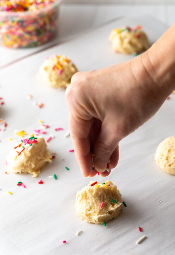 Hand adding colorful sprinkles to the tops of raw cookies balls spread out on a baking sheet.