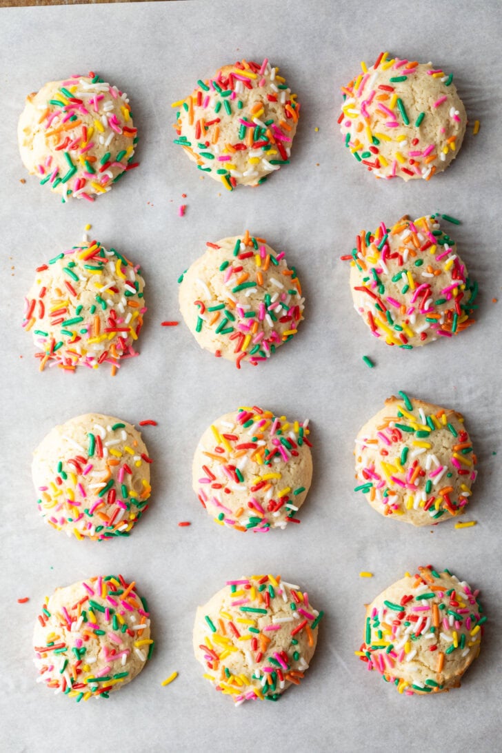 Top down view 12 cream cheese cookies on a baking sheet, each topped with sprinkles.