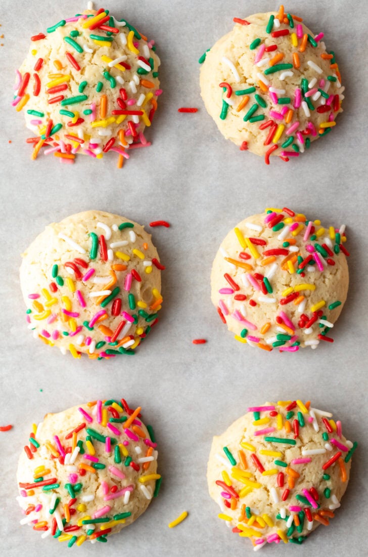 Top down view 6 cream cheese cookies on a baking sheet, each topped with sprinkles.