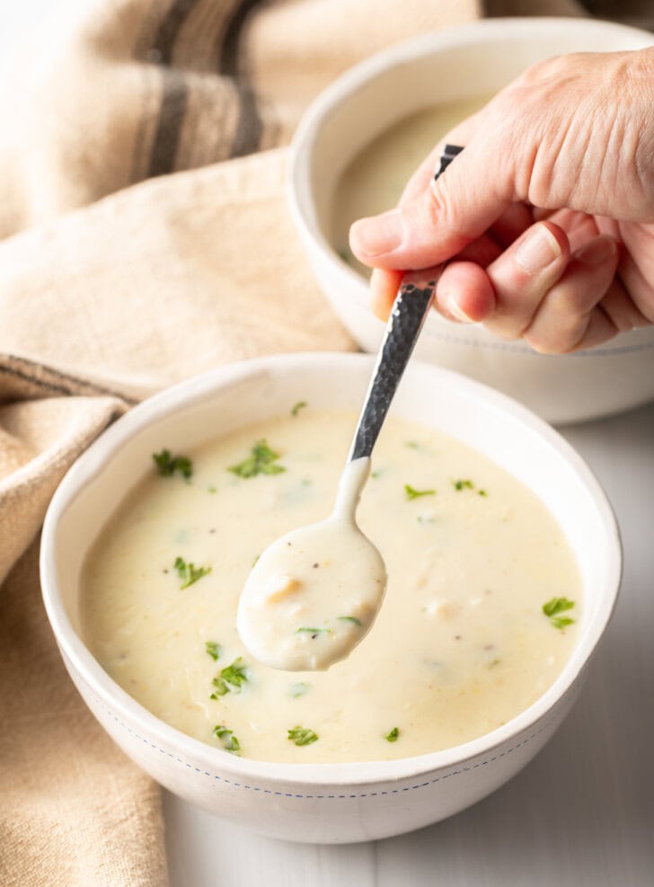 Top down view white bowl with creamy chicken soup garnished with fresh parsley. A hand with a metal spoon has a spoonful showing to camera.