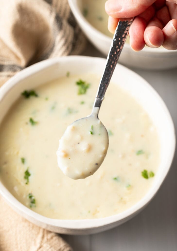 Top down view white bowl with creamy chicken soup garnished with fresh parsley. A metal spoon has a spoonful showing to camera.