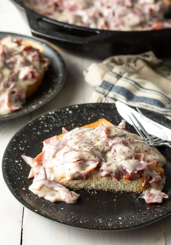 Creamed chipped beef on top of toast that has been cut in half on a black plate. 