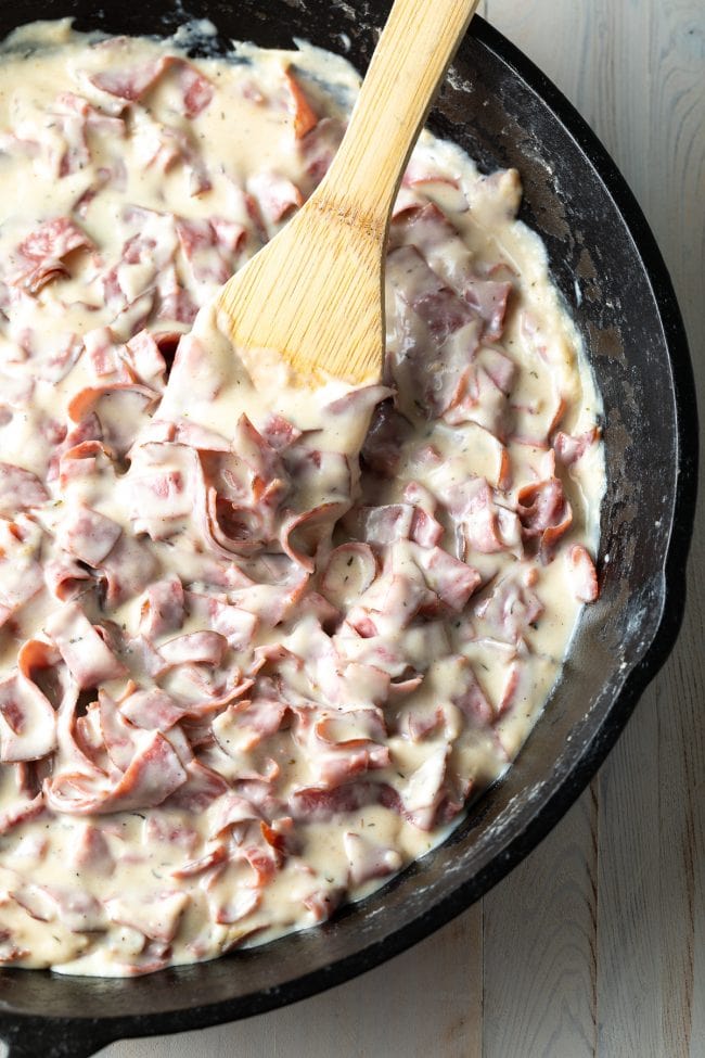 Chipped beef gravy from scratch being cooked in a cast iron skillet with a wooden spoon.