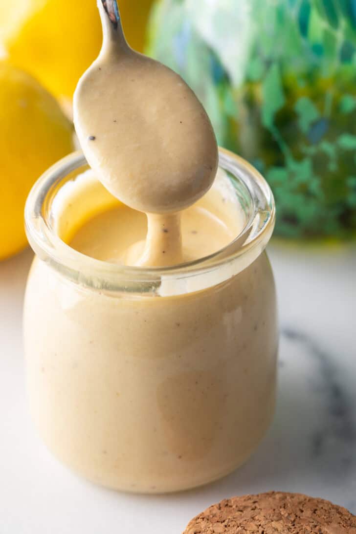 Spoon of dressing being held above a jar of tahini dressing.