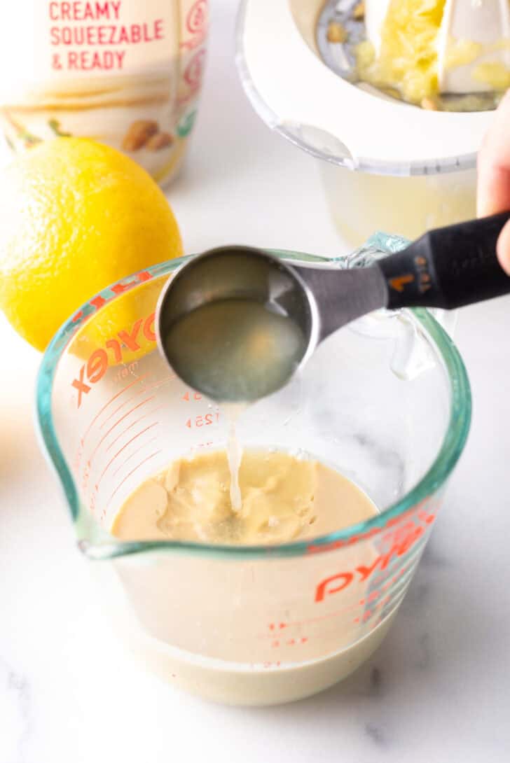 Hand adding lemon juice from a small measuring spoon into a glass measuring jar with tahini paste.