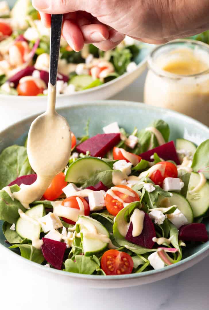 Hand with a spoon drizzling tahini dressing onto a greek salad with cucumbers, mixed greens, tomatoes, feta cheese cubes.