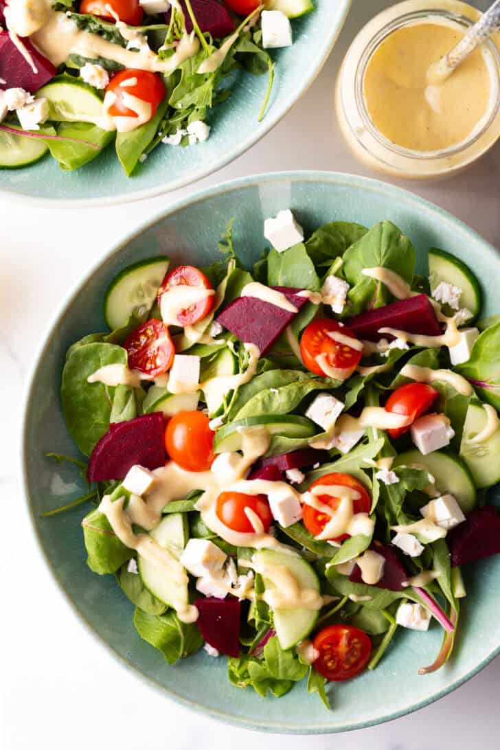 Top down view greek salad with cucumbers, mixed greens, tomatoes, feta cheese cubes, and a drizzle of pale yellow dressing.