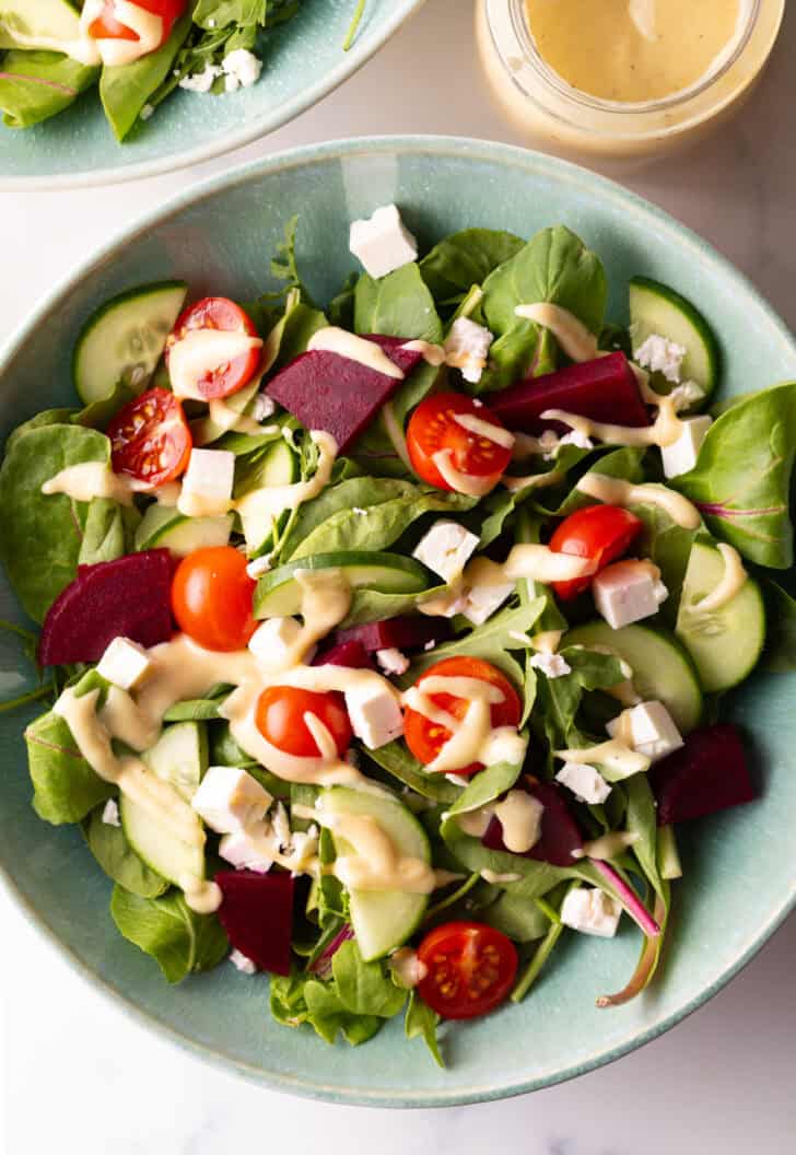 Top down view greek salad with cucumbers, mixed greens, tomatoes, feta cheese cubes, and a drizzle of pale yellow dressing.