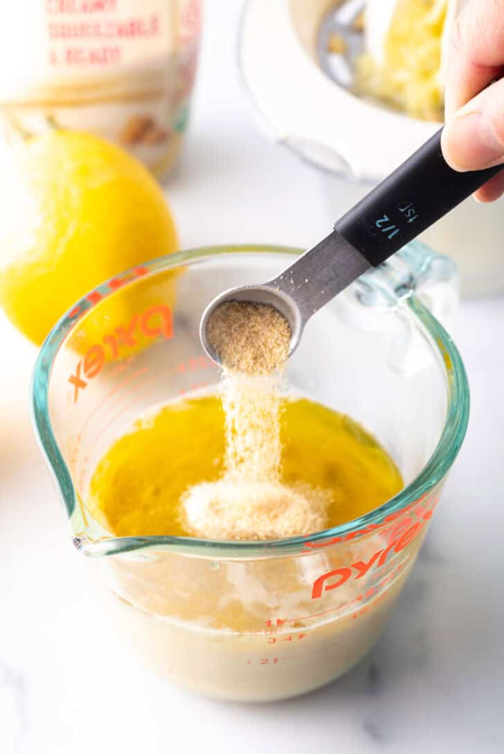 Hand adding spices from a small measuring spoon into a glass measuring jar with tahini paste.